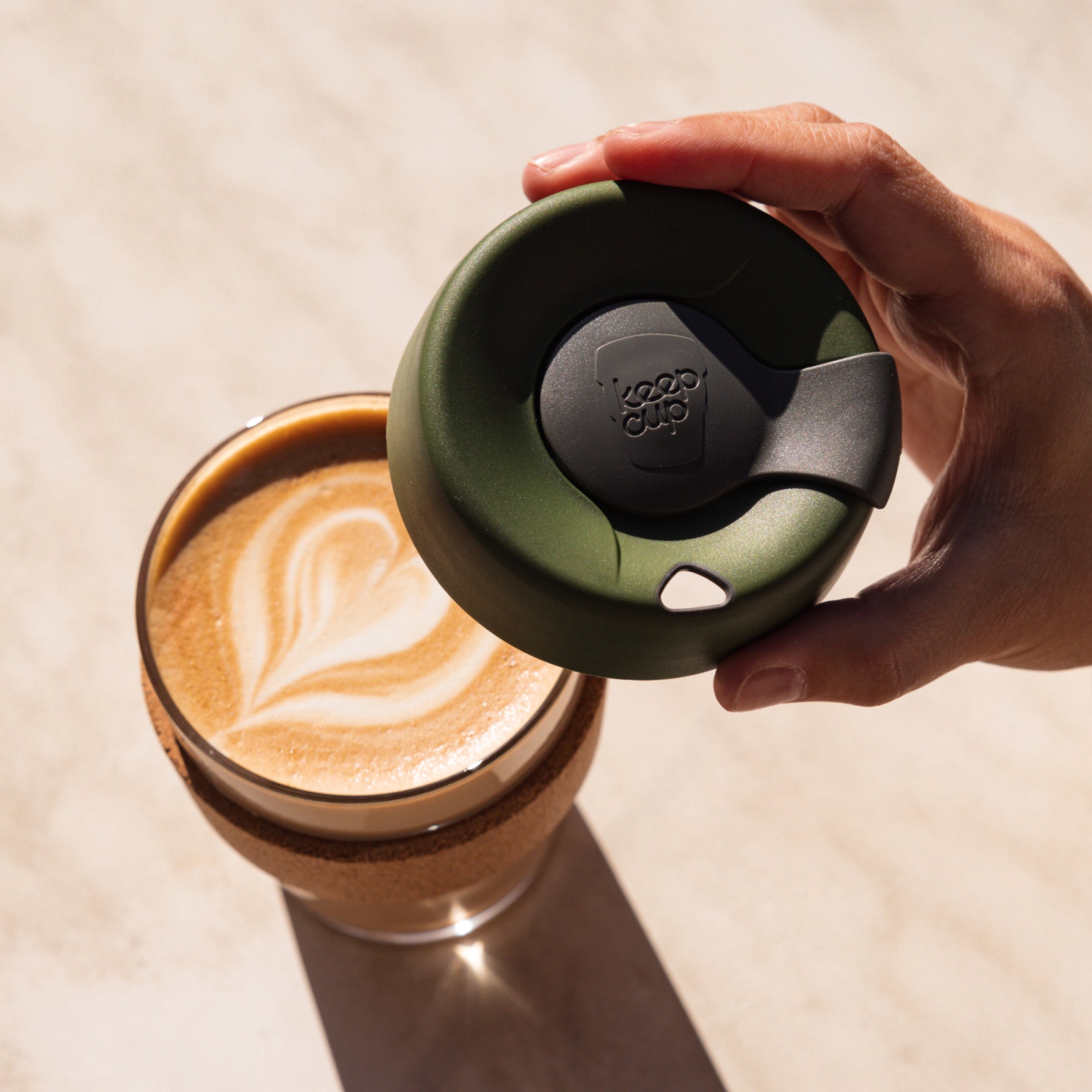 Hand holding a green coffee cup lid over a glass of coffee with latte art.
