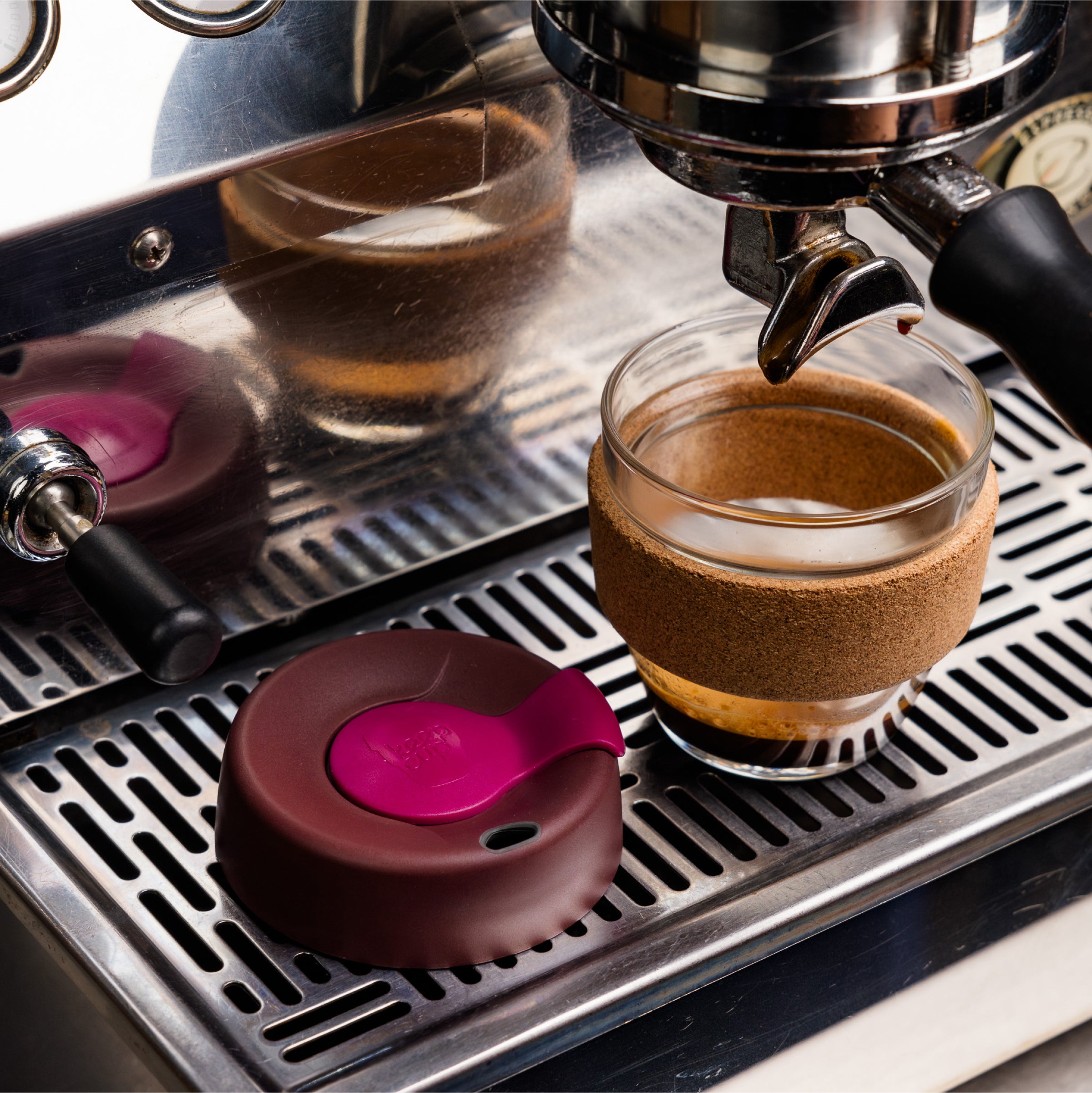 KeepCup Brew Cork S 8oz in Nutmeg colour sits beneath an espresso machine group head, with its burgundy and pink lid resting beside it on the stainless steel drip tray, showcasing the glass cup with natural cork band sleeve.
