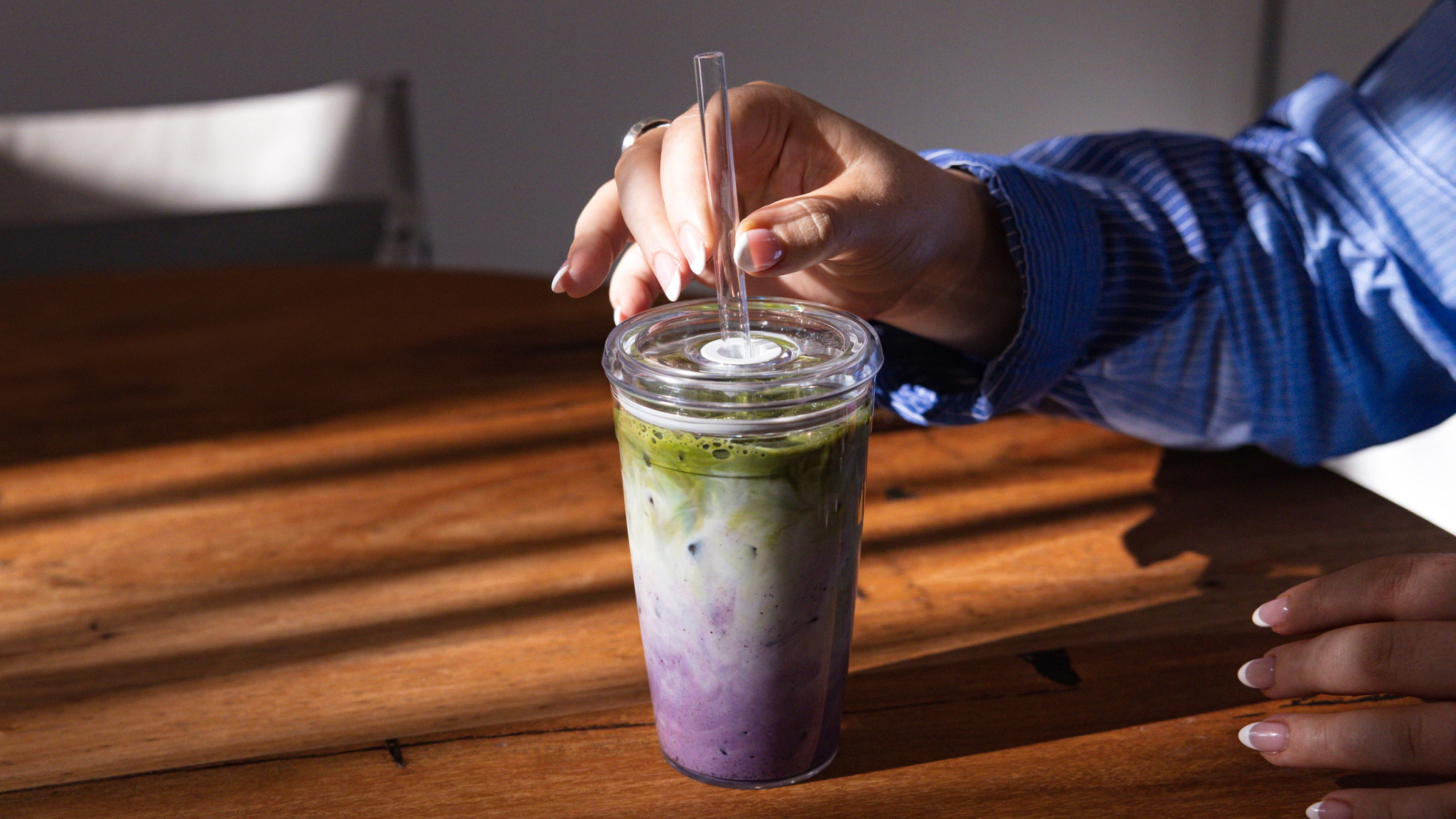 Person stirring layered green and purple smoothie with clear glass straw in reusable cup on wooden table