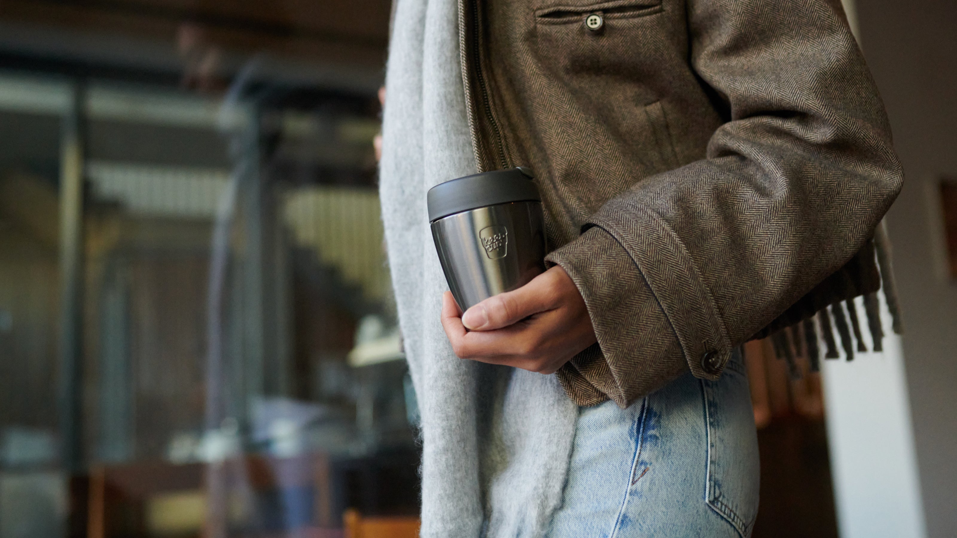Person holding black and silver insulated travel mug with logo, wearing brown herringbone jacket and grey scarf