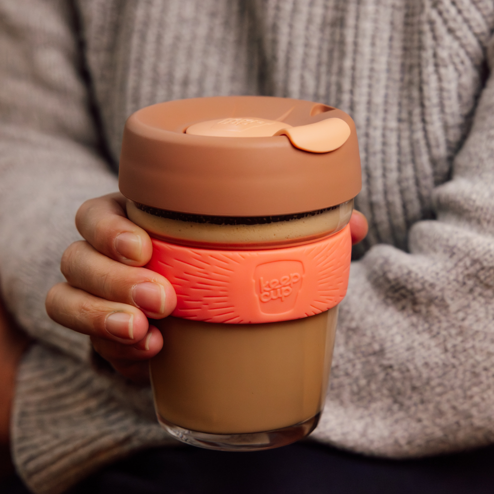 Person holding a glass KeepCup reusable coffee cup with a terracotta lid and sleeve.