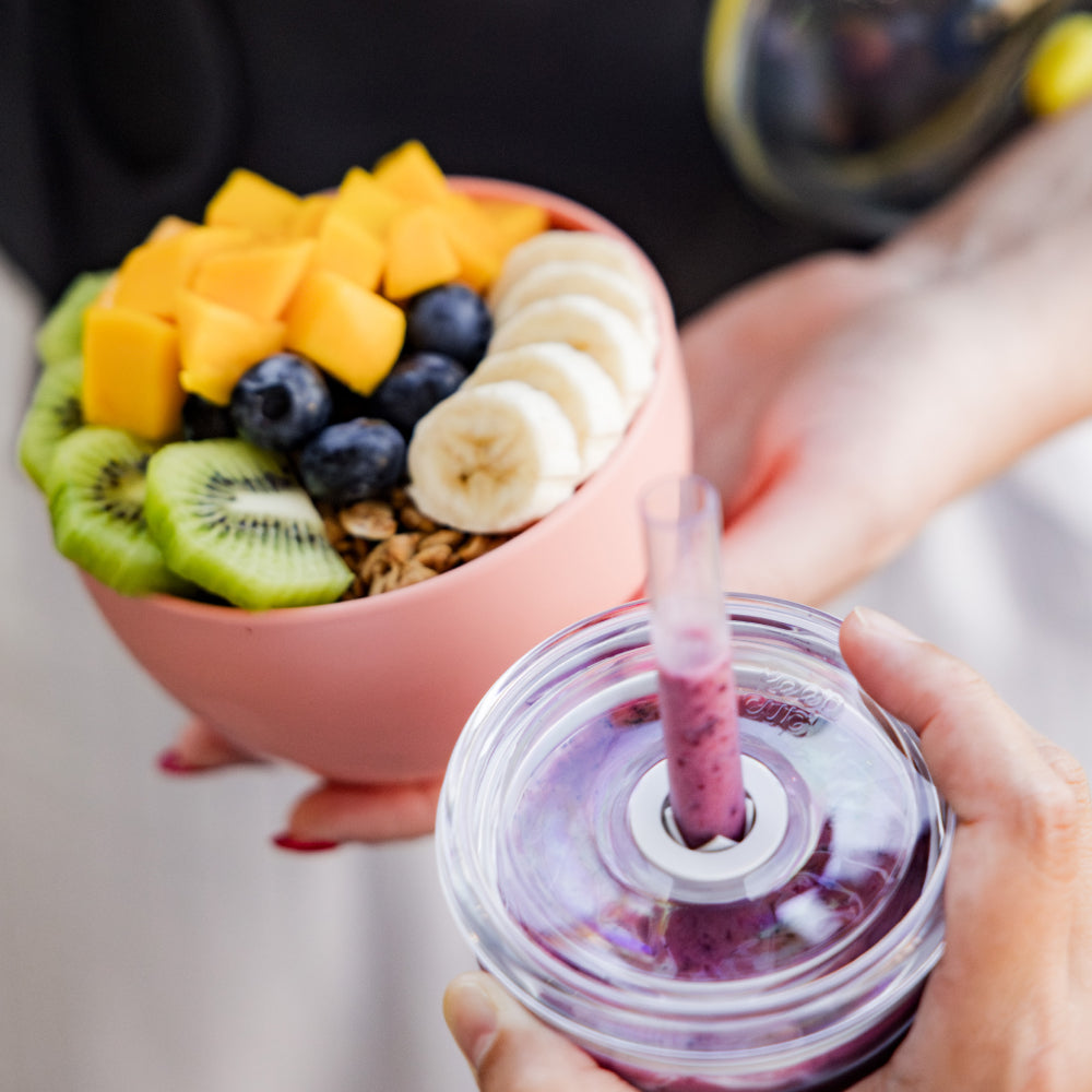 Person holding a purple smoothie in reusable cold cup with a straw, next to a bowl of fruit