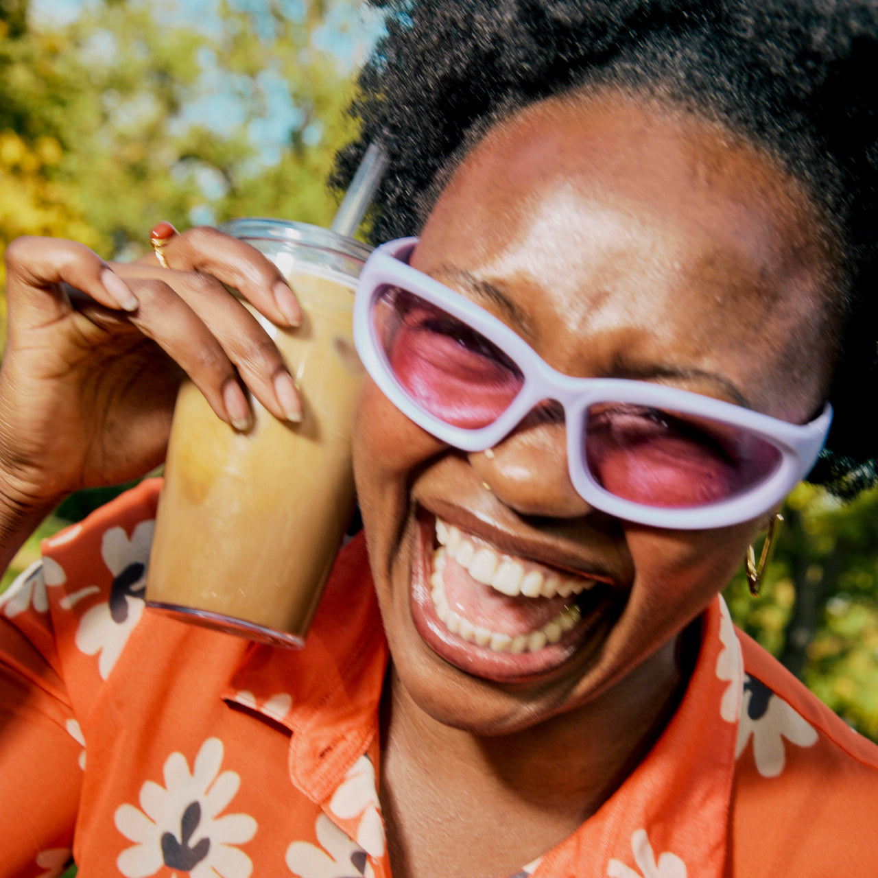 Person wearing pink sunglasses and an orange floral shirt, holding a drink outdoors.