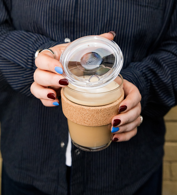 A person holding a reusable glass Brew Cork Quicksip with a cork sleeve and clear lid, shown in a close-up lifestyle shot.