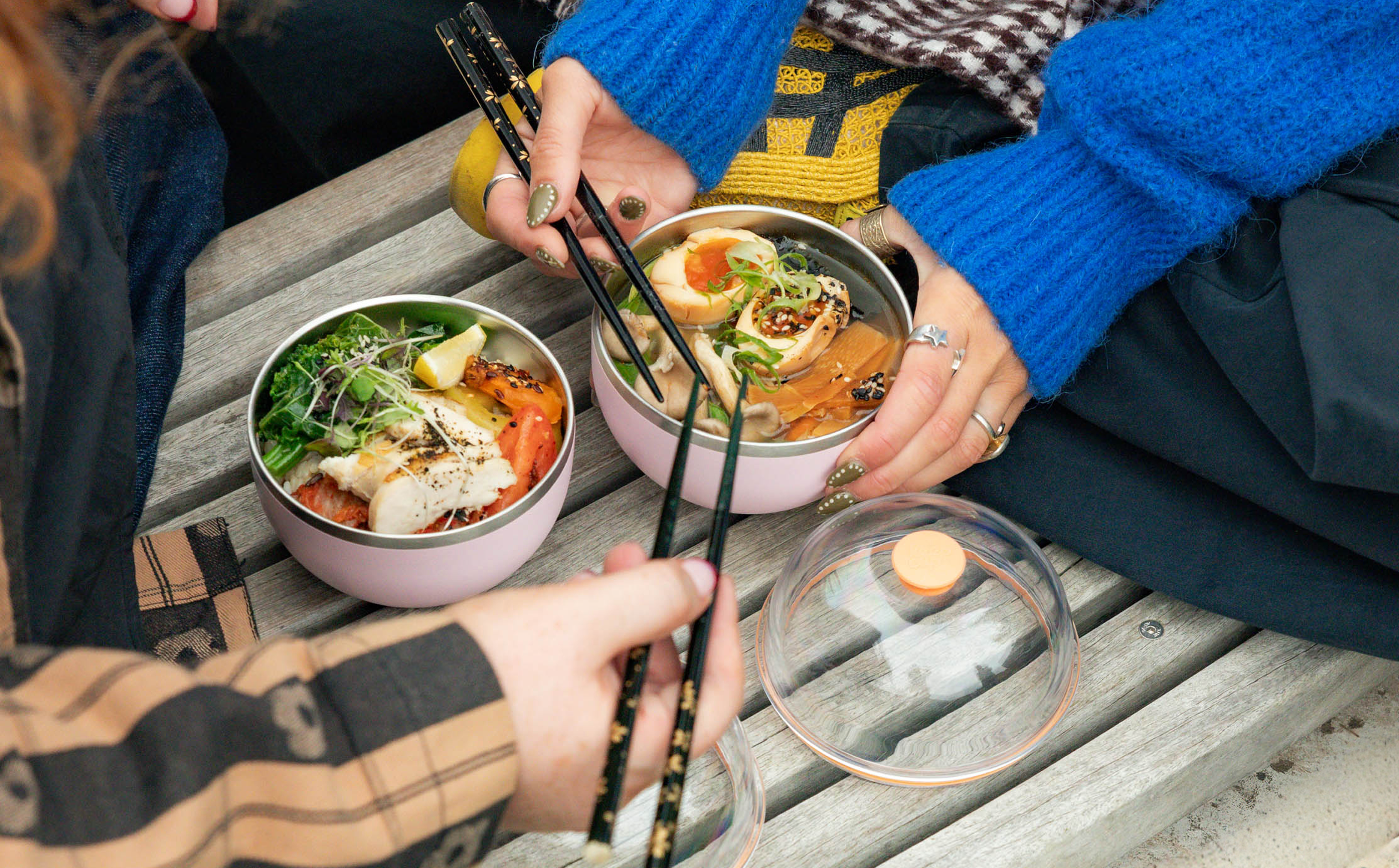 Two people enjoying a meal together with Go Bowl Luxe filled with food and chopsticks on a wooden bench.