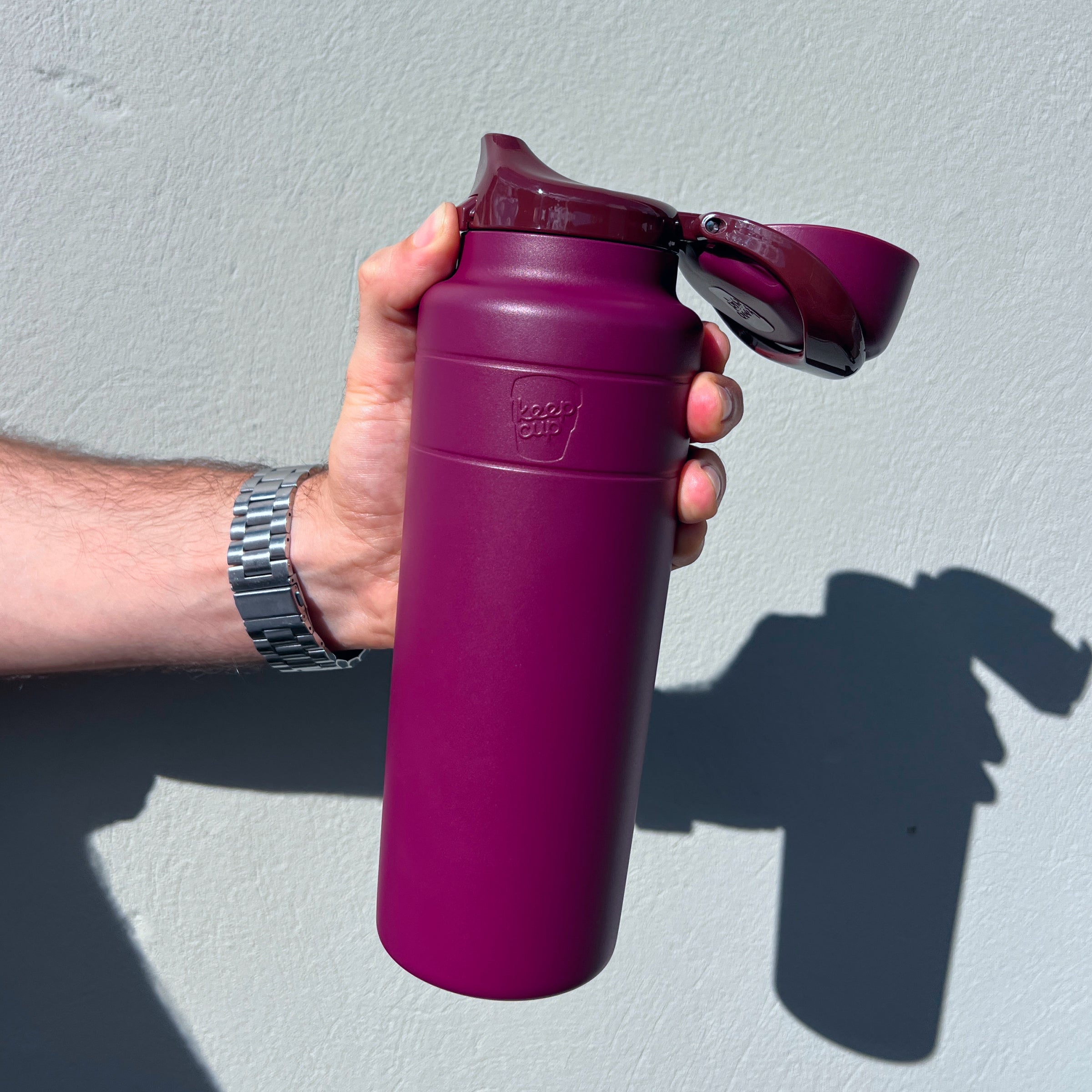 A hand holding the Ora Bottle 700ml in Mulberry, showing the open flip-top lid with the deep magenta matte body and KeepCup logo embossed on the front, photographed against a light grey wall in bright sunlight casting a shadow.