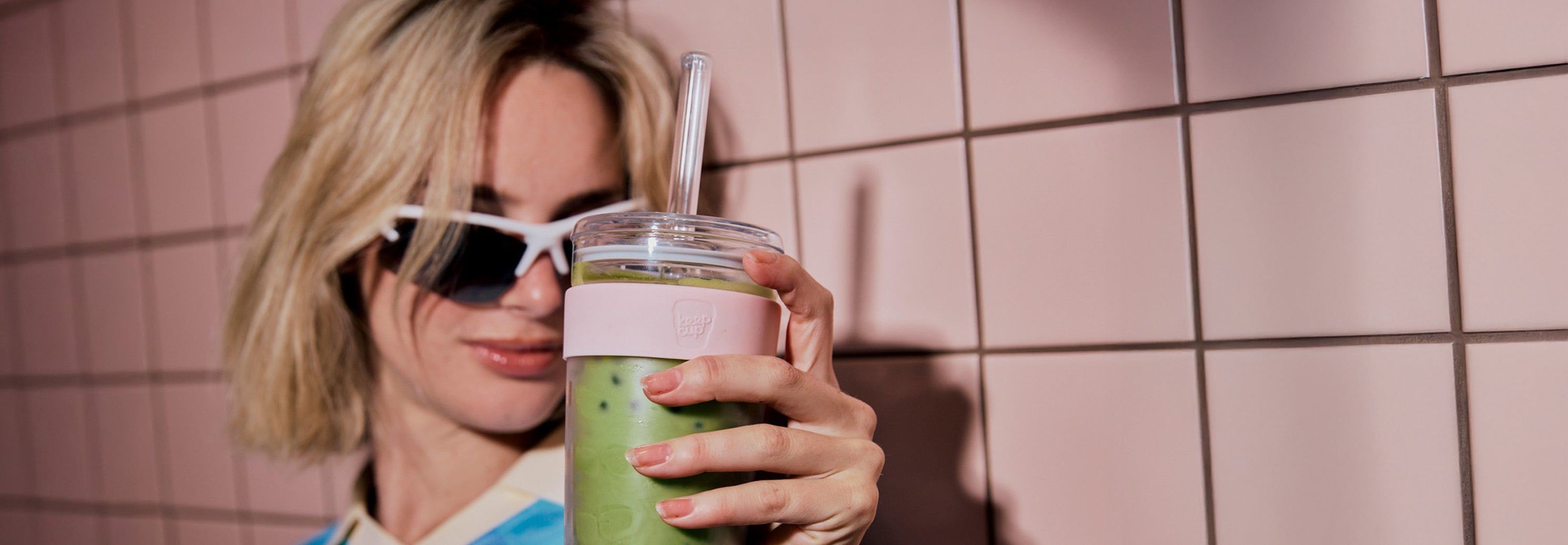 Woman holding a green smoothie in a clear cup with a straw against a tiled wall.