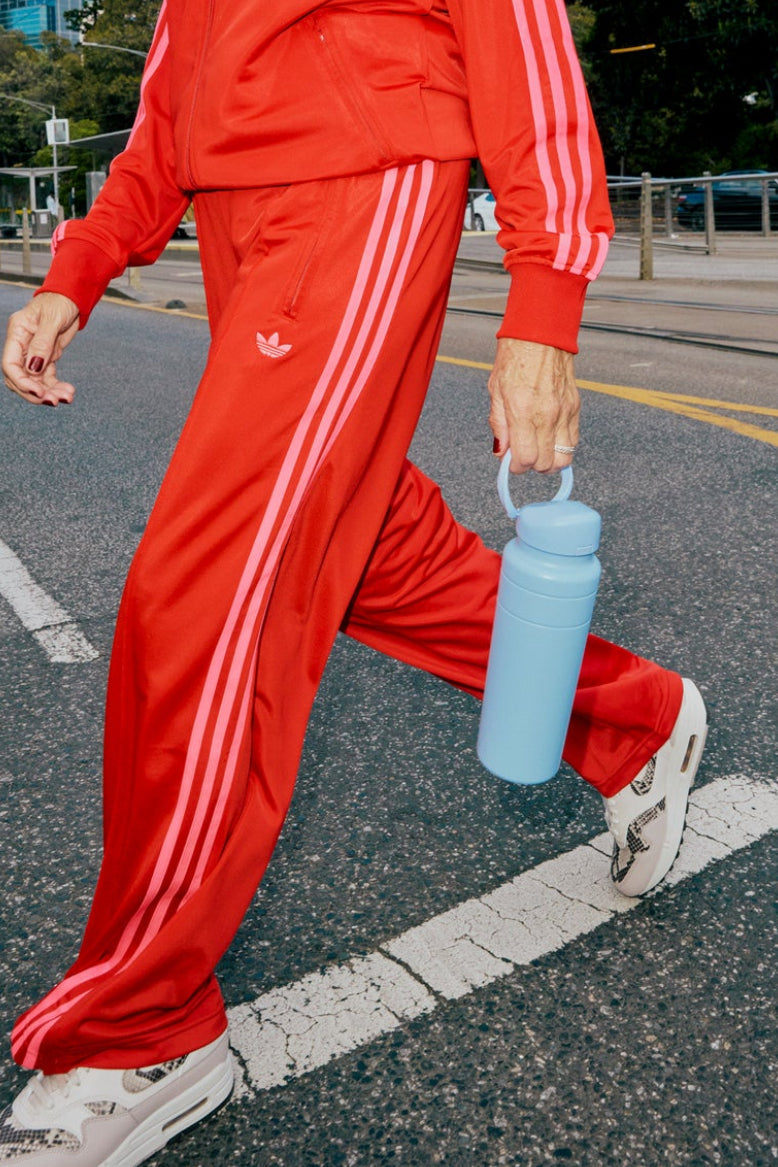 Person wearing a red Adidas tracksuit walking on a street with an orchid water bottle.