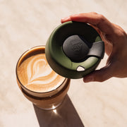 Hand holding a green coffee cup lid over a glass of coffee with latte art.
