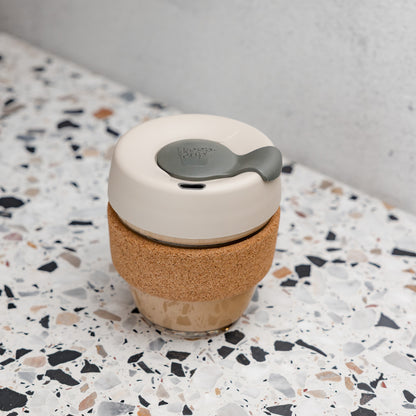 KeepCup Brew Cork XS 6oz in Filter colour sitting on a terrazzo surface, featuring a cream-coloured lid with grey plug, natural cork band, and glass cup filled with a milky coffee drink.
