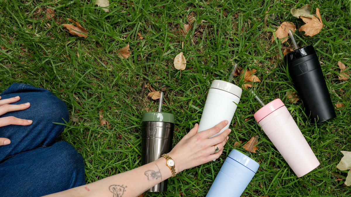 Collection of colourful reusable water bottles on grass - stainless steel, white, pink, blue and black bottles with person's tattooed hand reaching for one