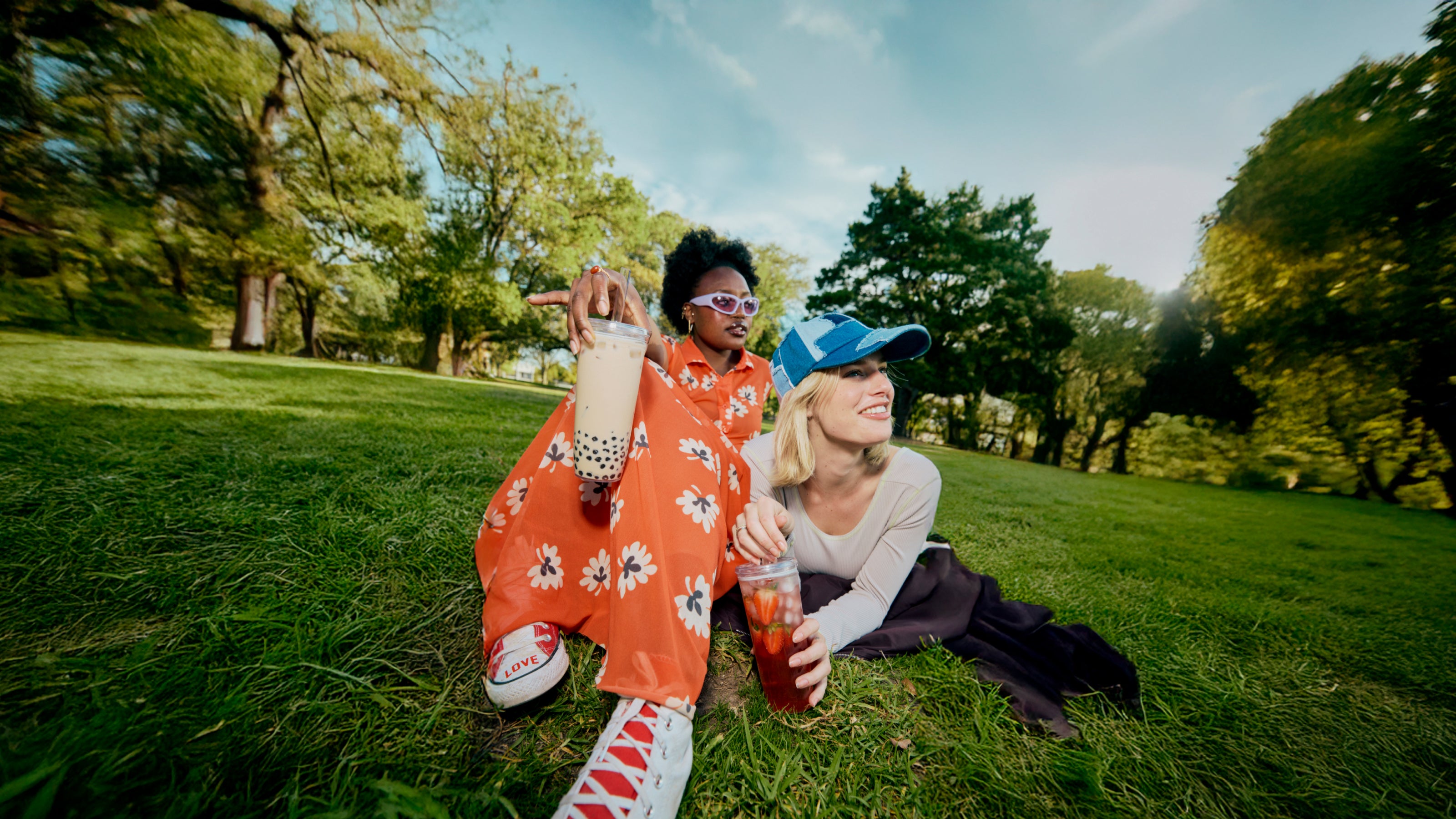 Two people sitting on grass in a park with trees and blue sky in the background