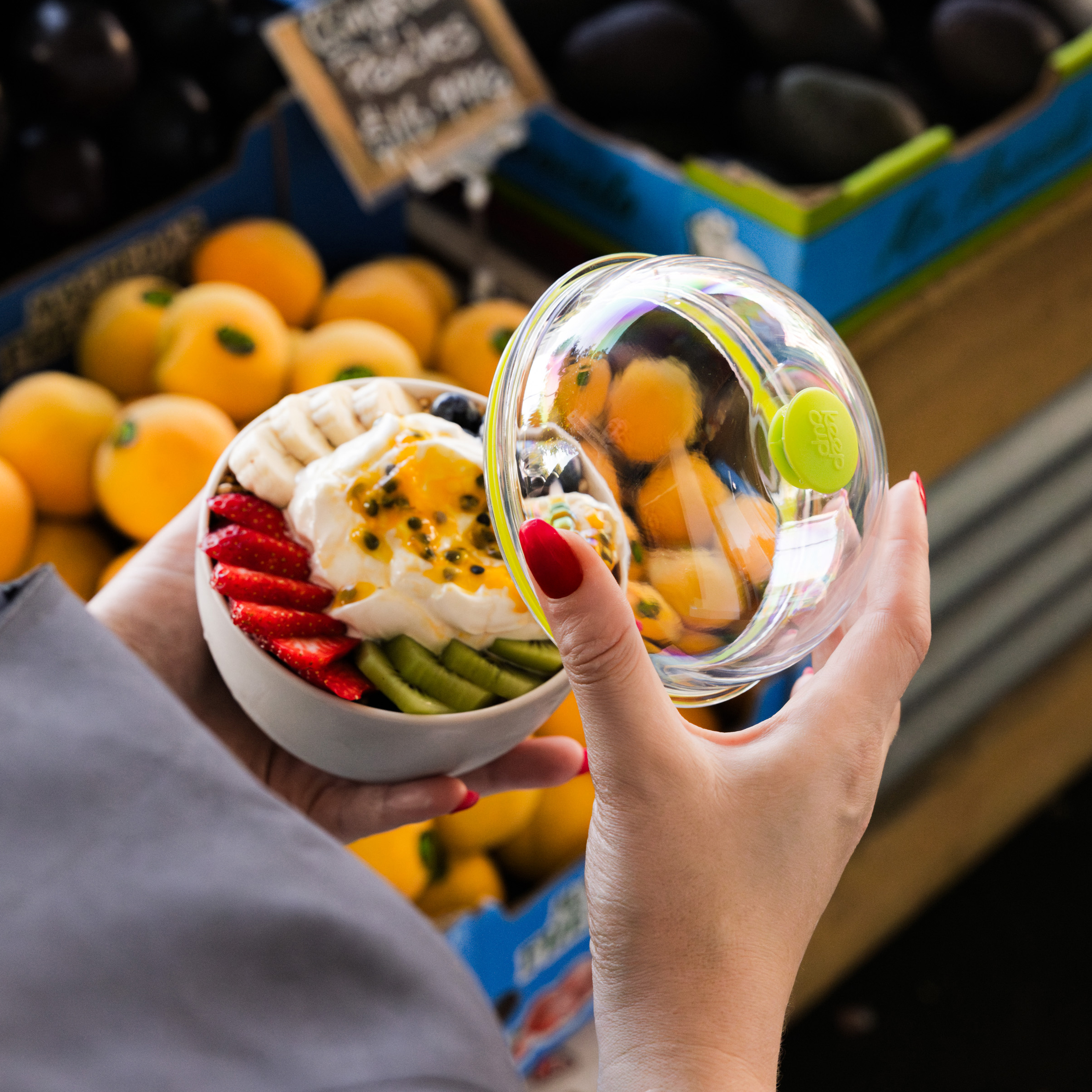 Clear glass bowl with green lid containing colourful fruit salad, held by hands in grocery store produce section