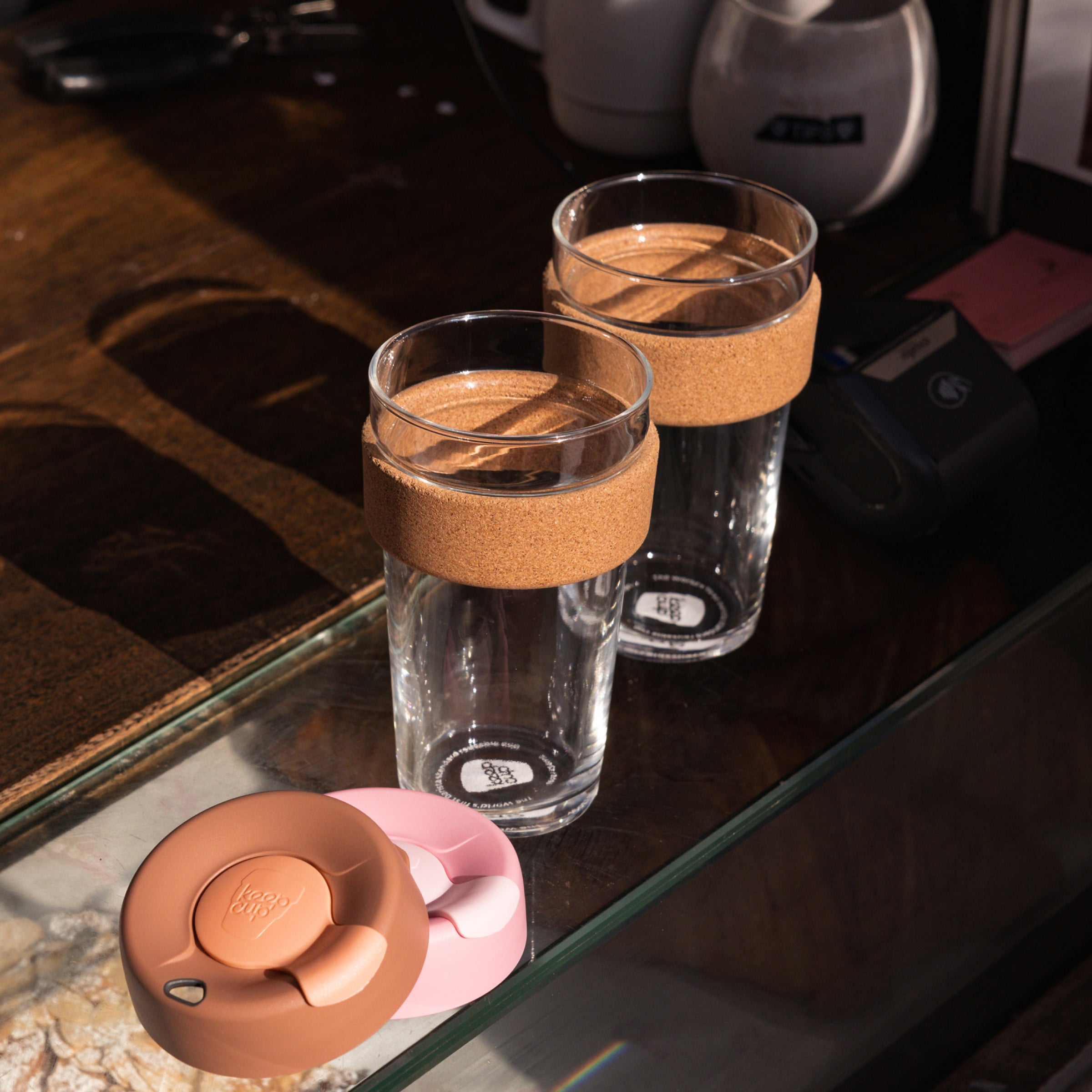 Two KeepCup glasses with brown cork bands and coloured lids on a reflective surface.