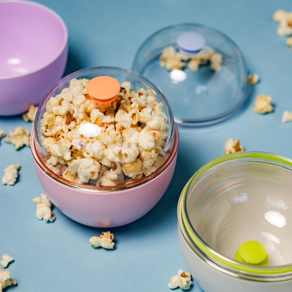 A pink Go Bowl Original M 550ml filled with popcorn, shown with its clear domed lid and orange plug, styled on a blue surface alongside lavender and green Go Bowl variations with scattered popcorn pieces.