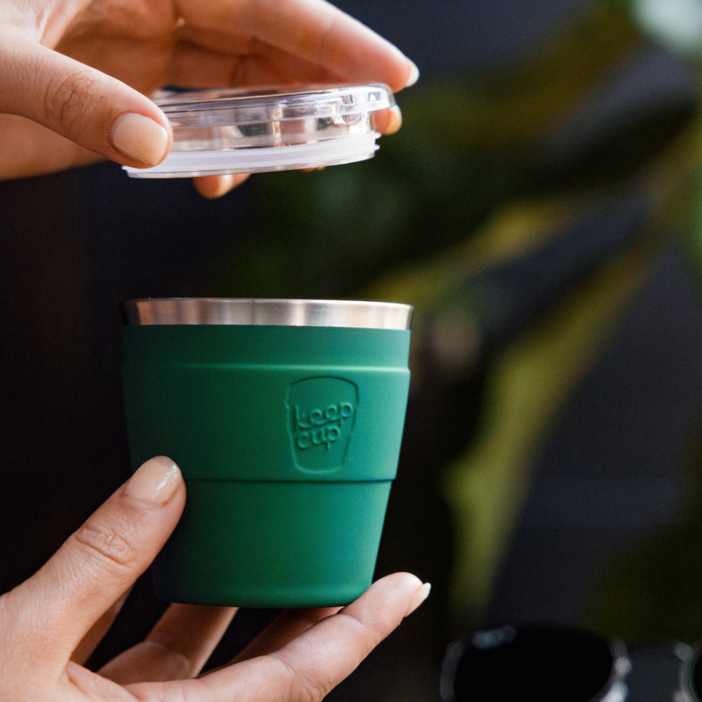 Hands holding a Thermal Quicksip XS 6oz reusable cup in Frost Green, with one hand lifting the clear plastic lid to show the stainless steel interior rim and matte green silicone band with embossed KeepCup logo.