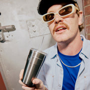Man wearing sunglasses and a cap, holding a silver tumbler with a straw.