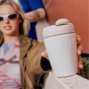 Person holding a white reusable coffee cup with 'keep cup' branding, blurred background