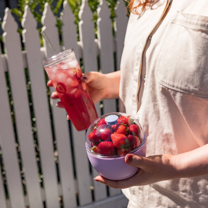 Person holding a pink drink with ice and a bowl of strawberries outdoors.