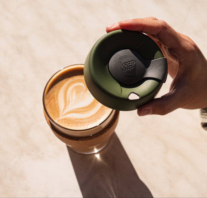 Hand holding sage green KeepCup reusable coffee cup with black lid above glass of latte with leaf art on beige surface
