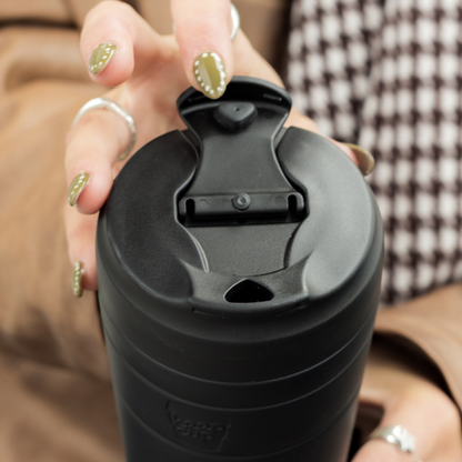A close-up of a person holding a black insulated KeepCup travel mug with a flip lid. The person's hands are visible, showing gold glitter nail polish and silver rings, with a brown coat and a brown-and-white checkered shirt in the background.