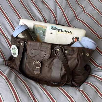 A brown leather shoulder bag filled with a laptop, two light blue KeepCup bottle and travel mug, resting on a gray and red striped bedsheet background.