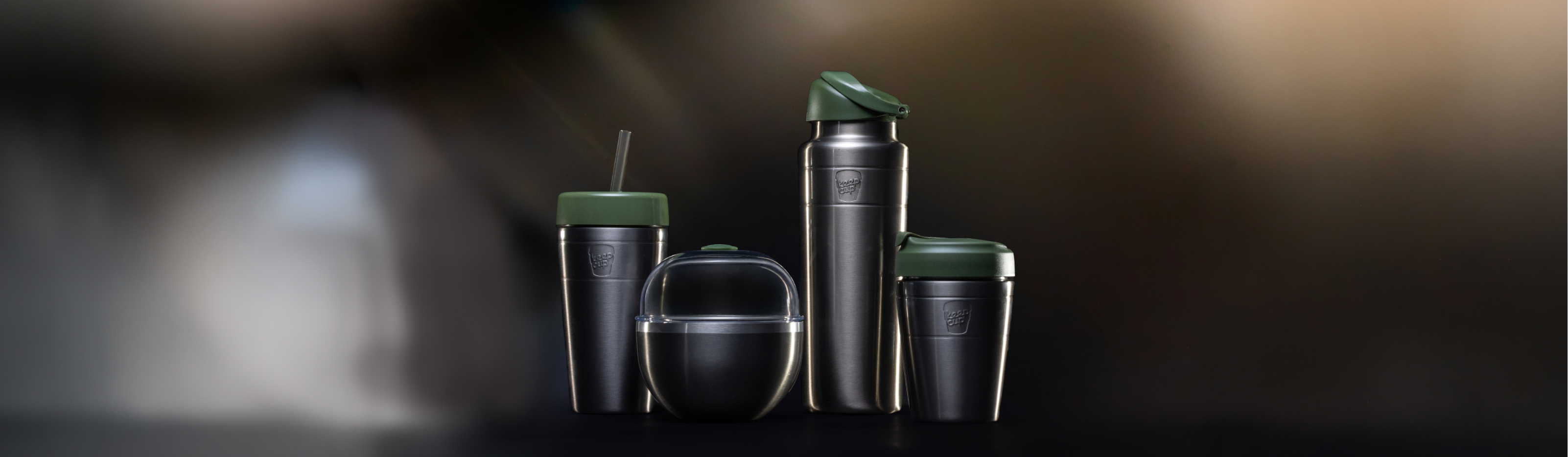 Four stainless steel travel containers with dark pine lids are arranged on a dark surface against a blurred warm brown background. The set includes a  water bottle, tall tumbler with straw, a small coffee tumbler, and a rounded container.