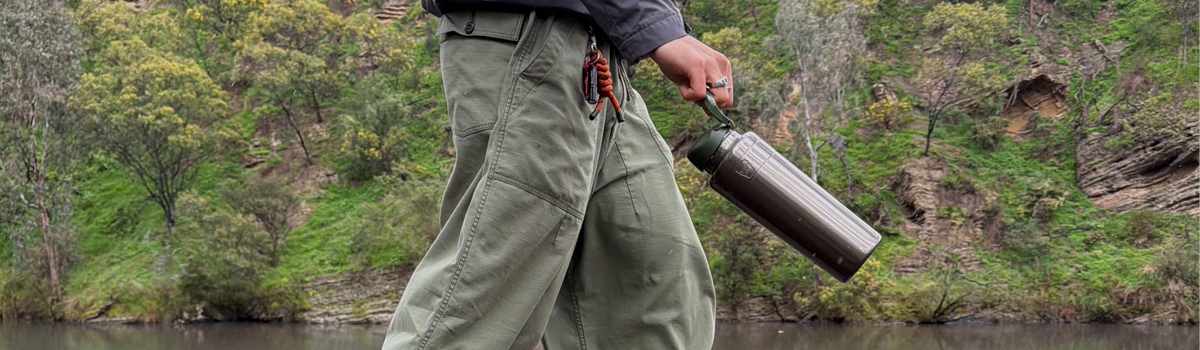 A person outdoors by a river or lake, wearing olive green cargo pants and holding a Nitro PIne Ora bottle. Trees and rocky cliffs are visible in the background.