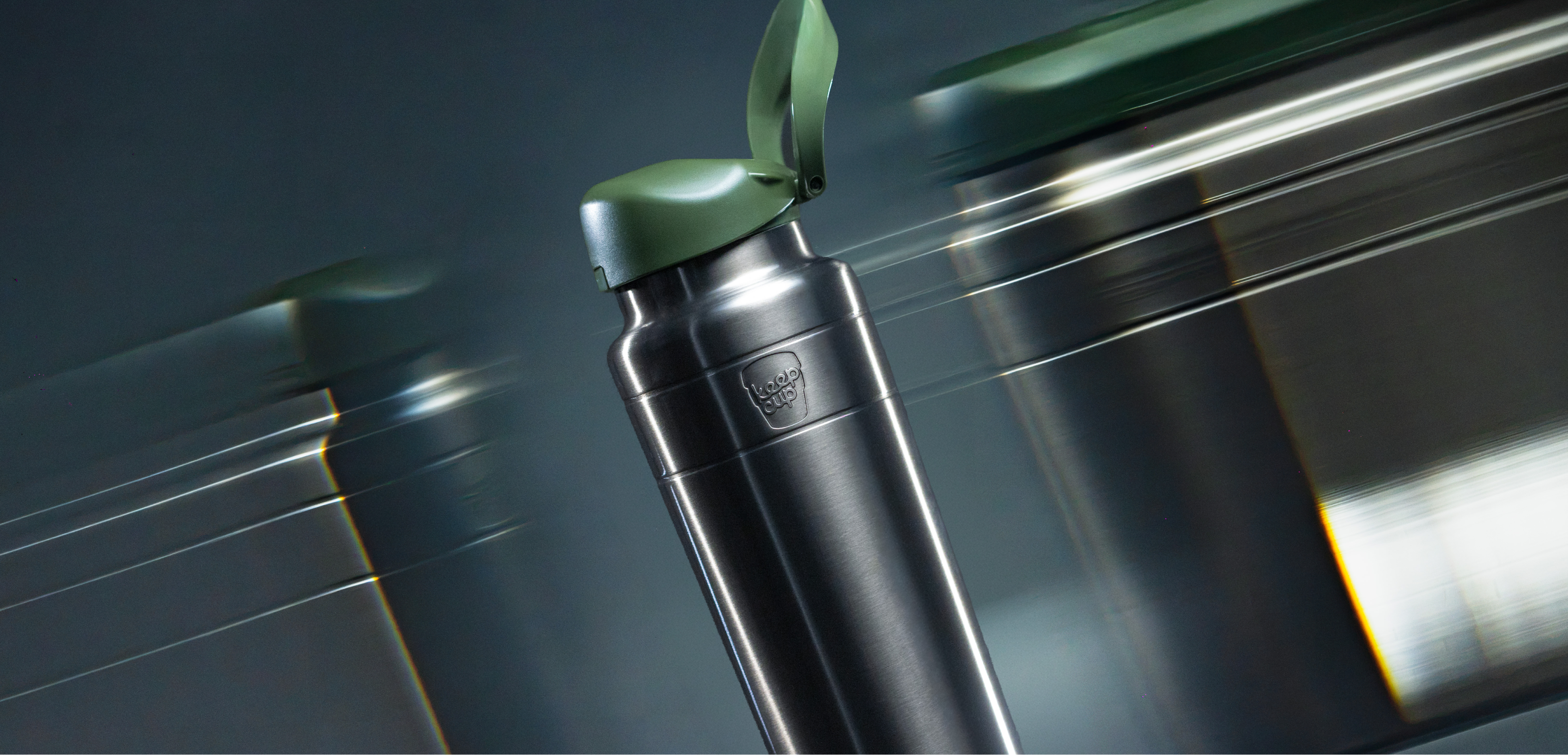 A sleek stainless steel Ora insulated water bottle in Nitro Pine with a dark green lid, shown in sharp focus with blurred motion effects of similar bottles in the background.