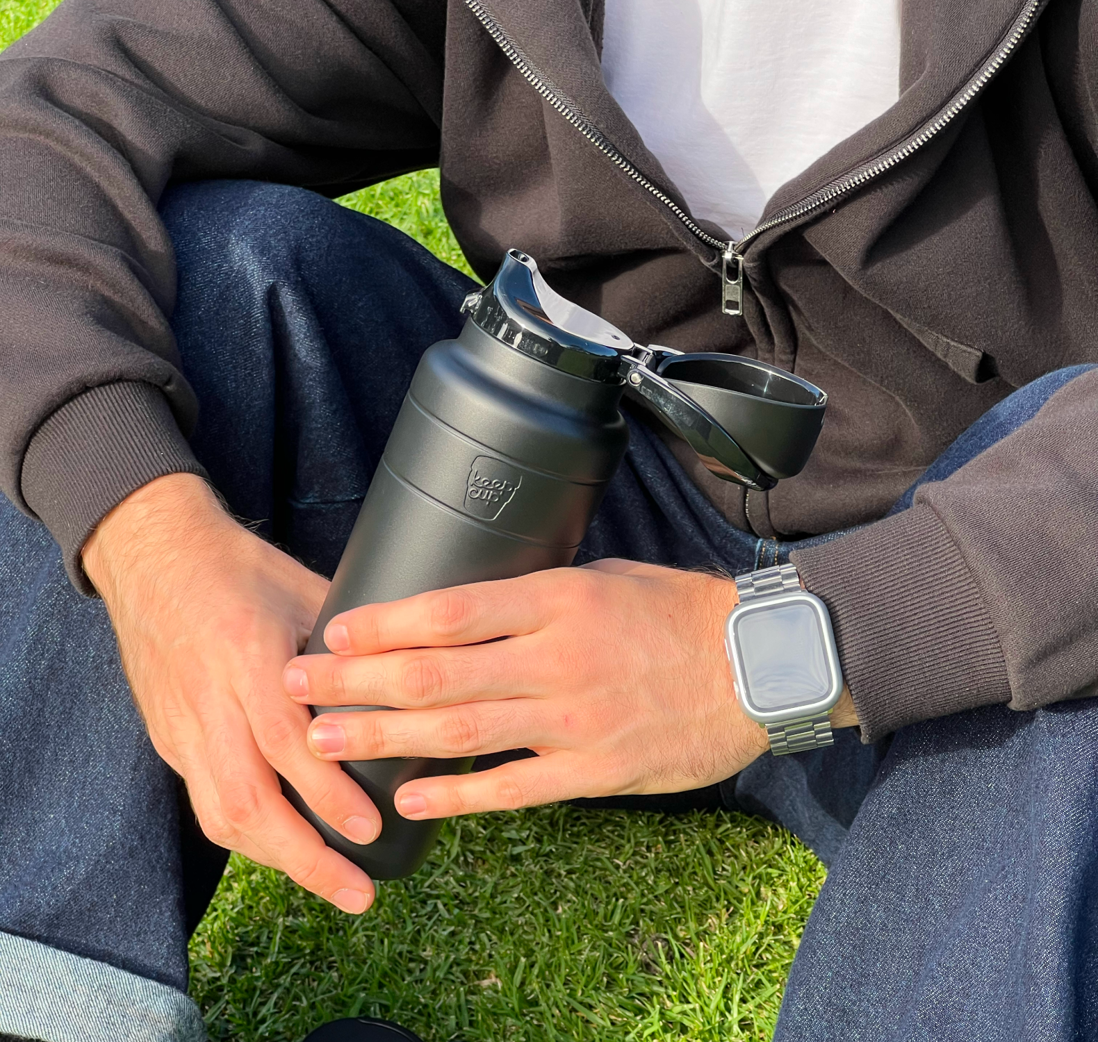 A person sitting outdoors on grass holds an Ora insulated water bottle in Blackwith its lid open. The person is wearing a dark zip-up hoodie, blue jeans, and a silver watch.