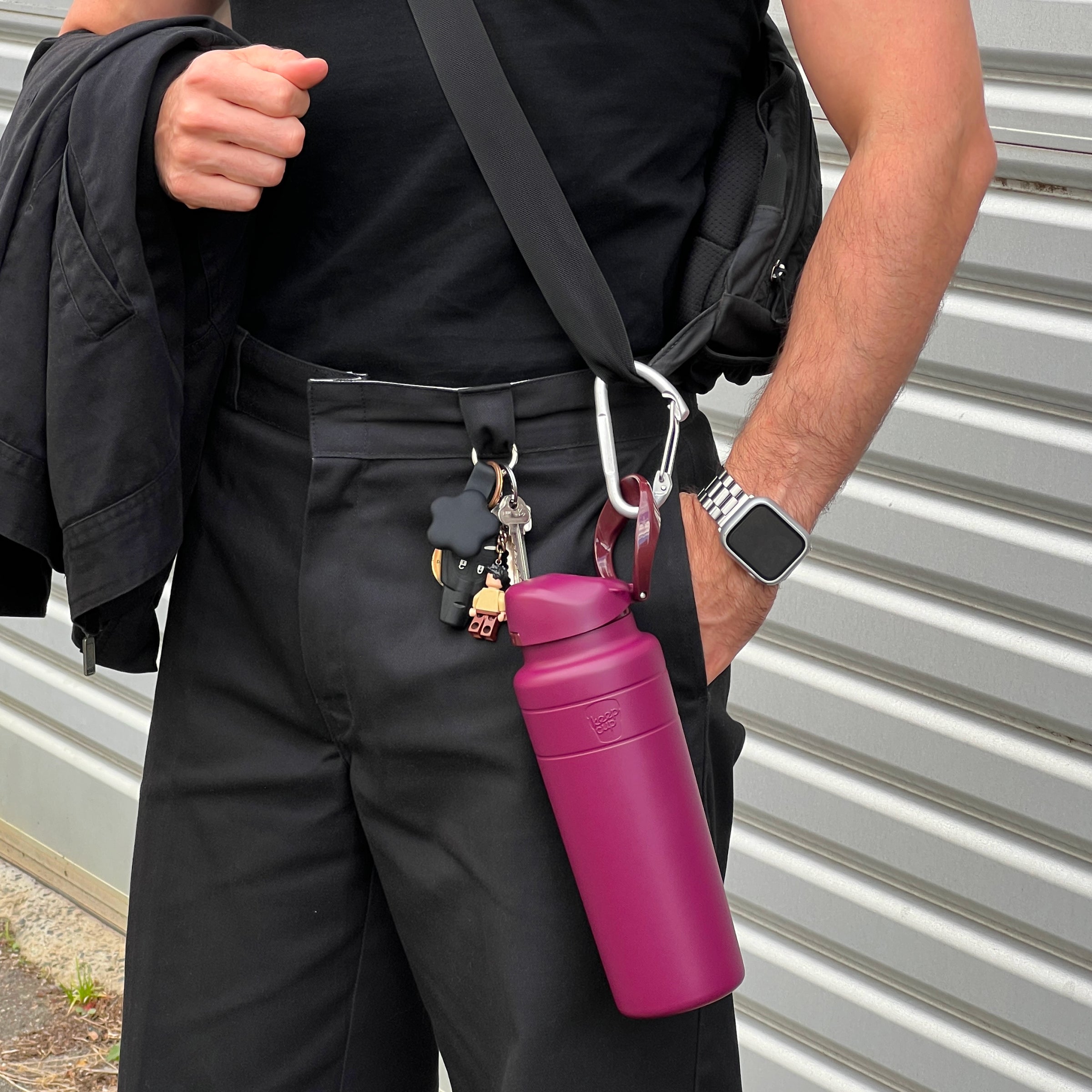 A person in black clothing carries the KeepCup Ora Bottle 700ml in Mulberry, clipped to their backpack strap with a carabiner, showcasing the bottle's built-in carry loop and deep magenta colour alongside keys and a small LEGO figure keychain.