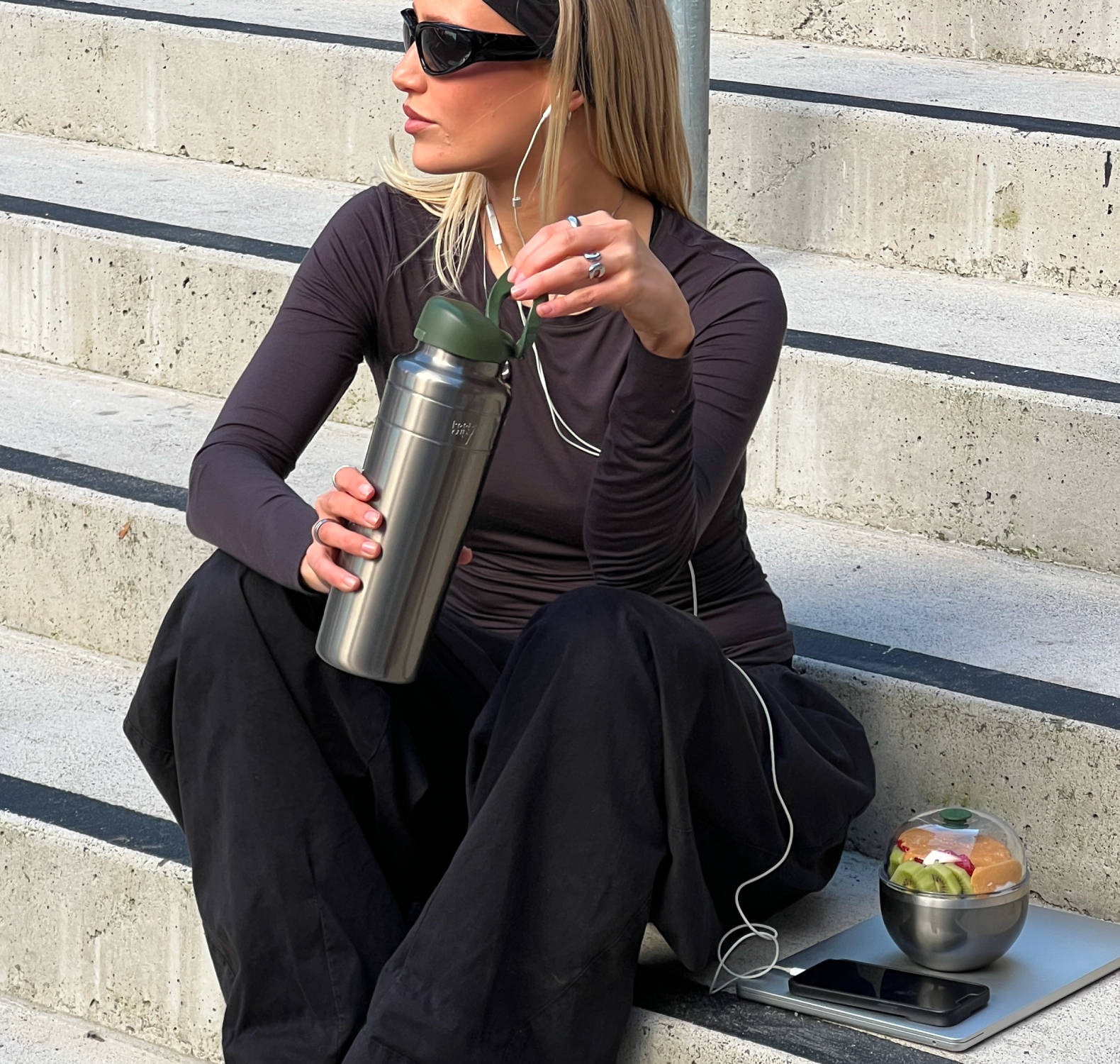 A woman sits on concrete outdoor steps wearing a fitted dark plum long-sleeve athletic top and black pants, holding an Ora insulated water bottle in Nitro Pine. She has white earphones in, insulated Go Bowl Luxe filled with fruit and a phone sit beside her on the steps.