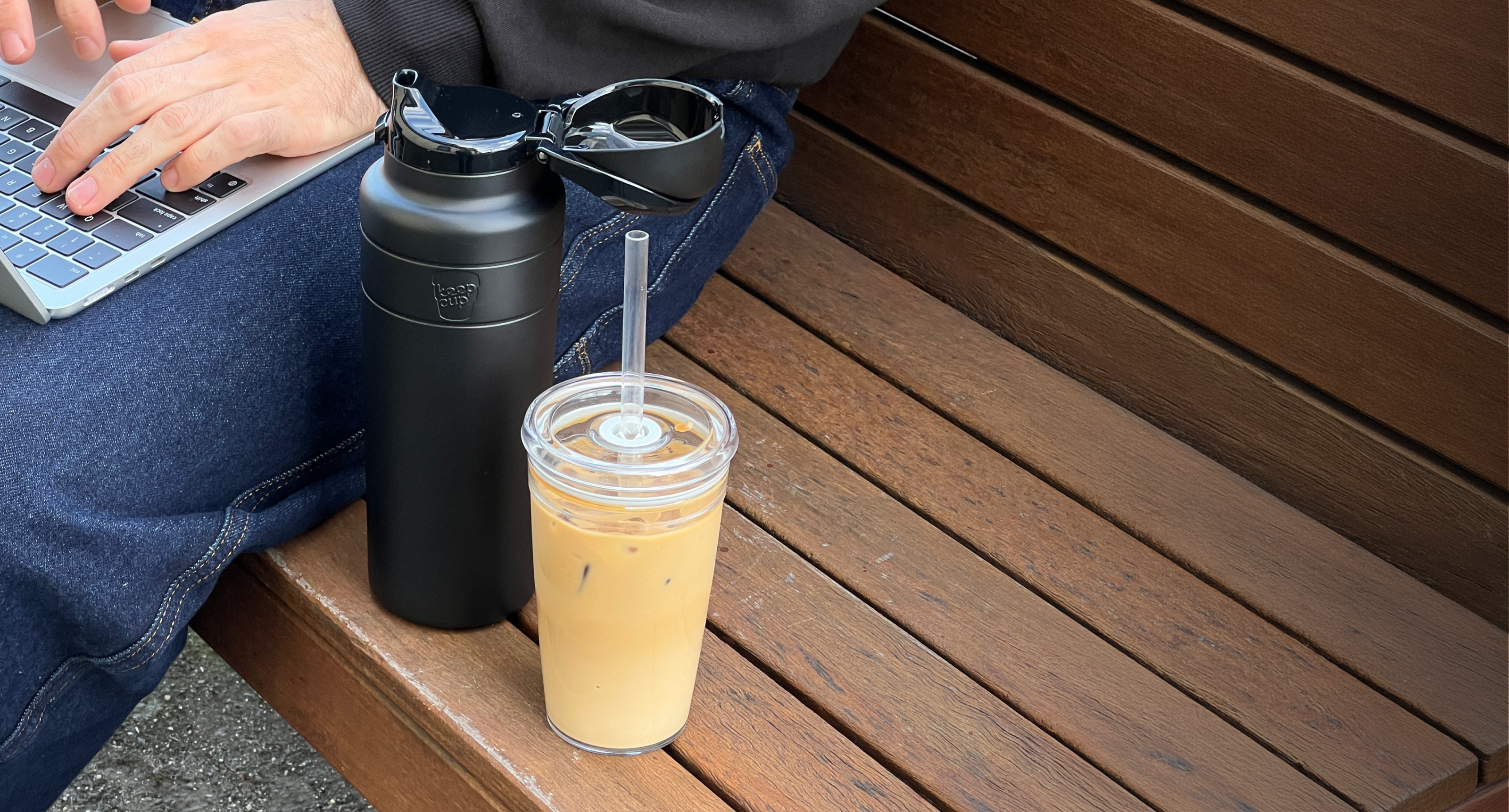 Black reusable water bottle with carry handle next to iced coffee drink on wooden outdoor table with laptop and denim jacket