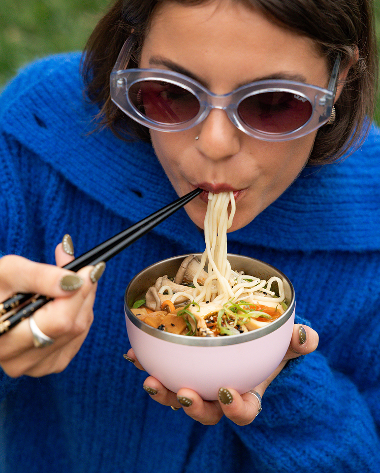 Person in blue sweater and sunglasses eating noodles from a bowl outdoors