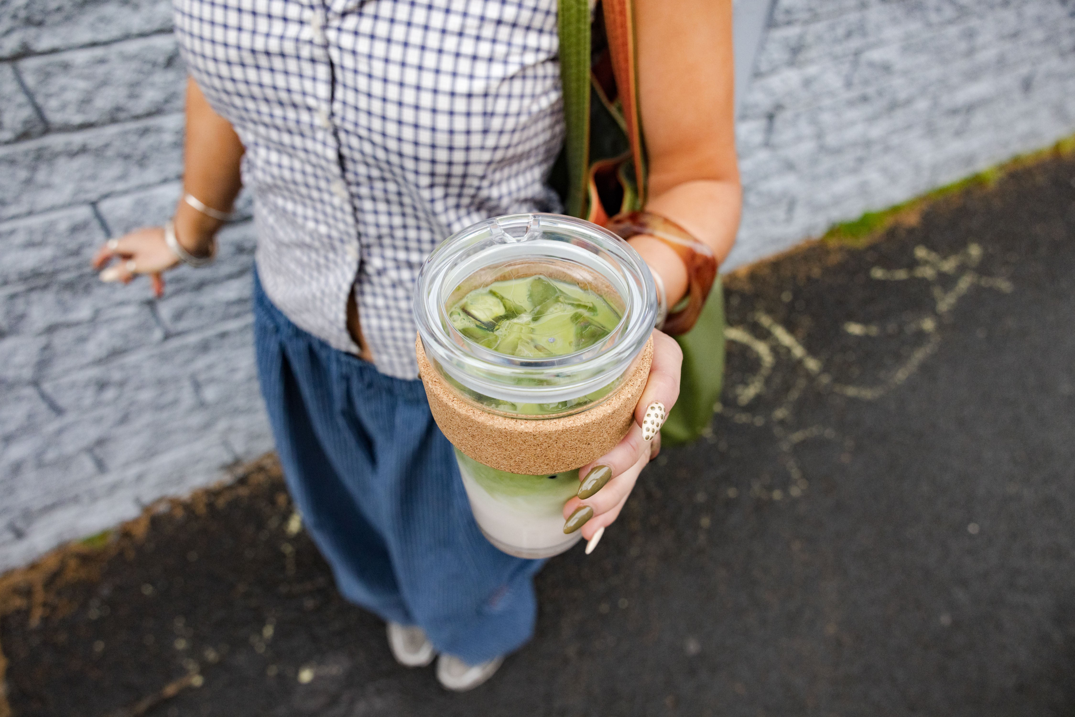 Person holding a glass jar with a green drink and straw sleeve on a street.