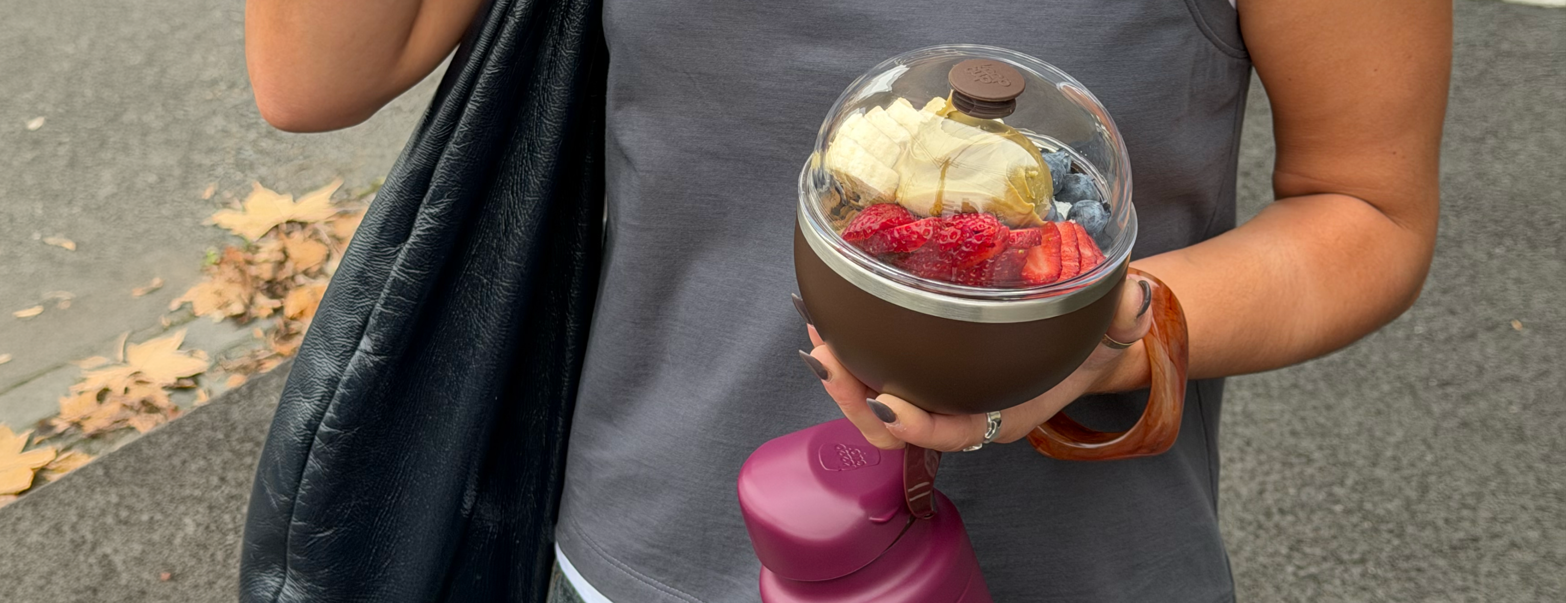 Person holding brown portable blender with clear dome lid containing sliced banana and strawberries, outdoors on pavement