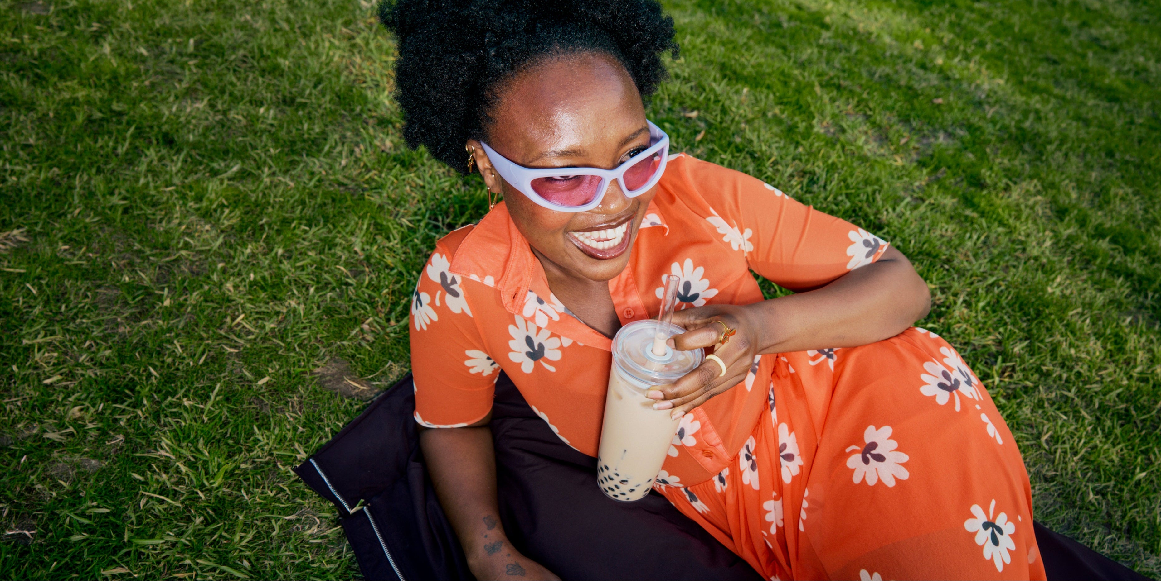 Woman in an orange floral dress sitting on grass holding a bottle.