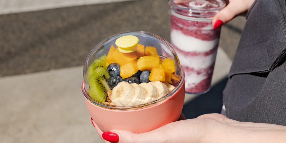 A woman crossing the street holding a KeepCup Cold Cup Original filled with a smoothie and KeepCup Go Bowl in Clay filled with fruit and acai.
